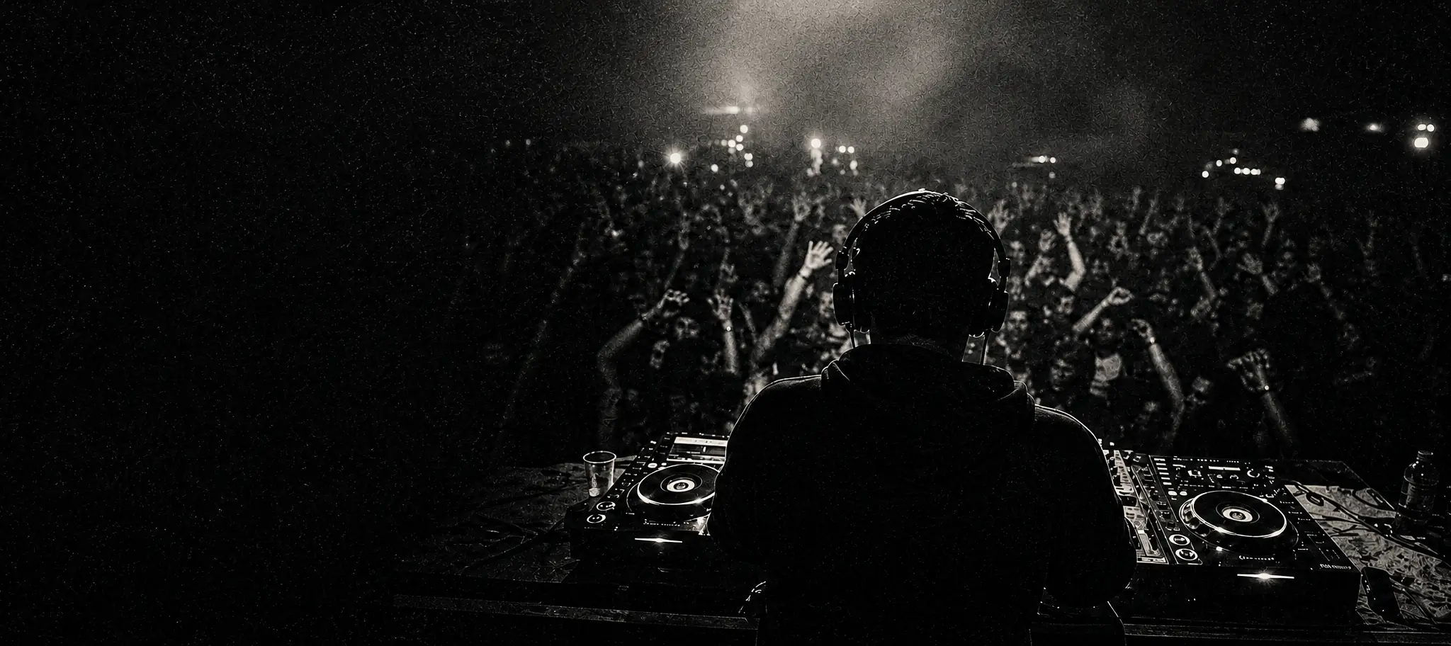A house music DJ seen from behind at the decks, facing a packed dark room with hands in the air